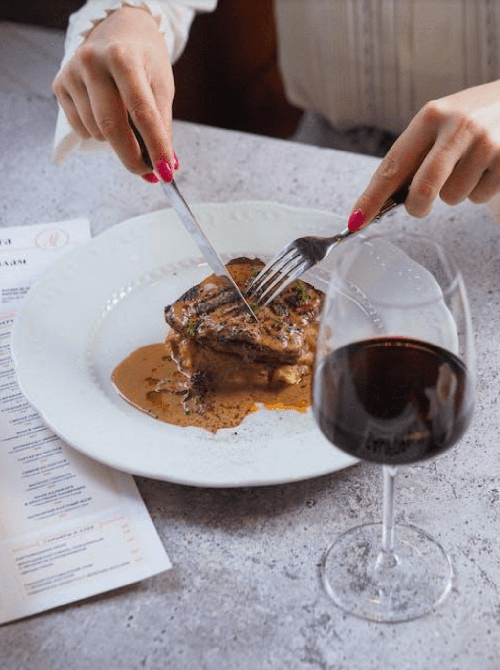 A person cutting through a steak with a glass of red wine next to the plate